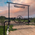 Ranch collection gate with custom iron branding logo with two roses beneath the logo with solar powered gate operator and with a custom overhead with ranch name hanging.